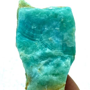 May include: A vibrant, raw gemstone with a striking turquoise hue. The stone displays a layered, textured surface with swirling patterns, and a rough, natural edge. The overall appearance is a blend of blue and green tones, with a hint of yellow at the edges.