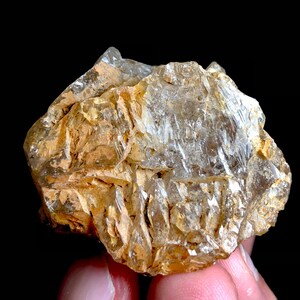 Puede incluir: Un grupo de cristales en bruto, sin cortar, con una mezcla de tonos claros y marrón dorado. La superficie rugosa muestra una estructura cristalina con bordes afilados y un aspecto natural. La muestra mineral se presenta sobre un fondo negro.