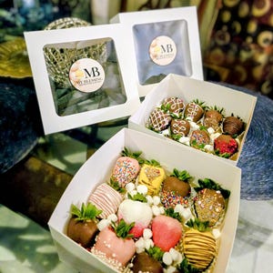 May include: White boxes with clear windows showcasing chocolate-covered strawberries. The strawberries are decorated with sprinkles, drizzles, and other toppings. The boxes have a circular sticker with the text "MB My Blessing Cakes & Desserts".