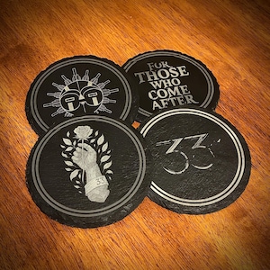 May include: Set of four black slate coasters with silver designs. One coaster features the text "FOR THOSE WHO COME AFTER." Another displays a hand holding a rose with the number "33." The other two coasters have geometric and numerical designs.