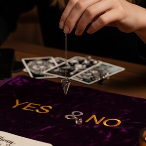 May include: A clear crystal pendulum suspended over a purple velvet cloth embroidered with "YES & NO" in gold. Tarot cards are visible in the background, suggesting a divination theme. The pendulum is held by a person.