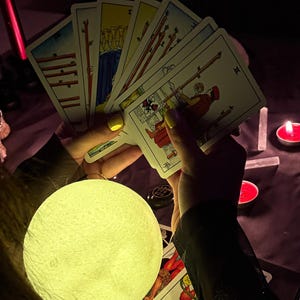 May include: A glowing moon-shaped lamp illuminates a spread of tarot cards held in hands with yellow nail polish. The cards feature colorful illustrations and are arranged in a fan shape. Red candles and crystals are in the background.