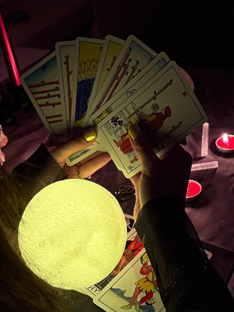 May include: A glowing moon-shaped lamp illuminates a spread of tarot cards held in hands with yellow nail polish. The cards feature colorful illustrations and are arranged in a fan shape. Red candles and crystals are in the background.