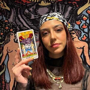 May include: A person holding a tarot card with the text "ASIKLAR". The card features a colorful illustration of figures and symbols. The person is wearing a patterned headscarf, a sheer black top, and multiple necklaces. A tapestry with figures is in the background.