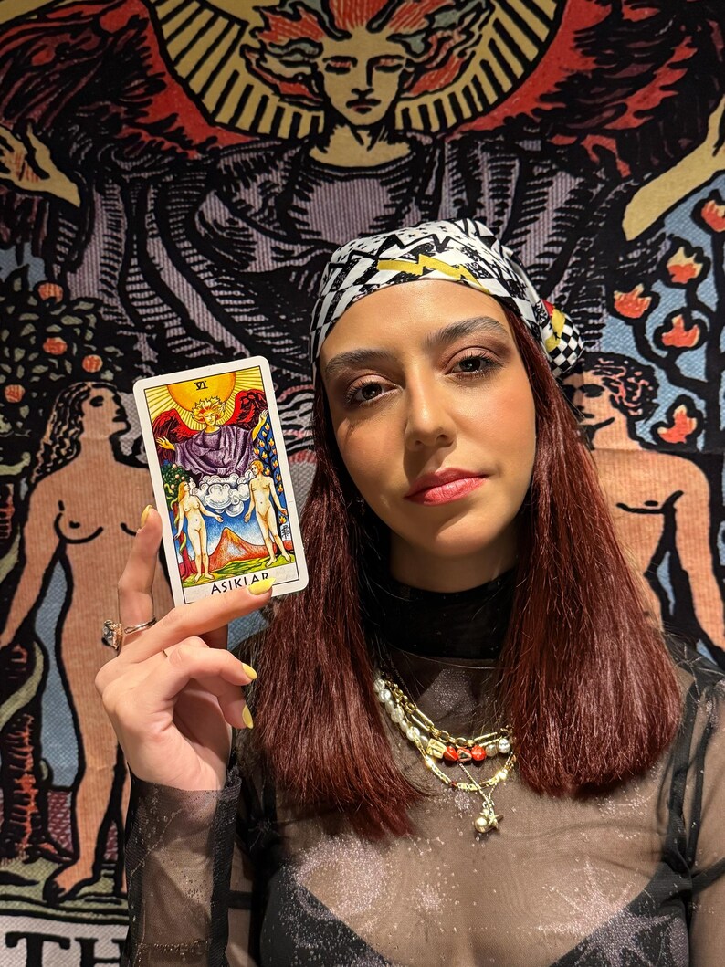 May include: A person holding a tarot card with the text "ASIKLAR". The card features a colorful illustration of figures and symbols. The person is wearing a patterned headscarf, a sheer black top, and multiple necklaces. A tapestry with figures is in the background.
