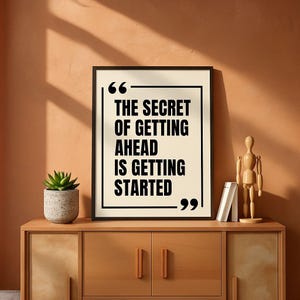 May include: A framed print with the quote "THE SECRET OF GETTING AHEAD IS GETTING STARTED" in black text. The print is displayed on a wooden cabinet with a succulent plant and a wooden artist's mannequin.