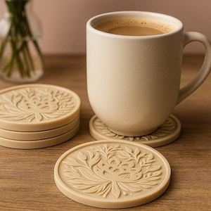 Op de afbeelding: Een beige keramische mok gevuld met koffie staat op een bijpassende onderzetter met een bloemenpatroon. Er liggen meerdere extra onderzetters in de buurt. De onderzetters en de mok staan op een houten oppervlak. Een vaas met groene stelen staat op de achtergrond.