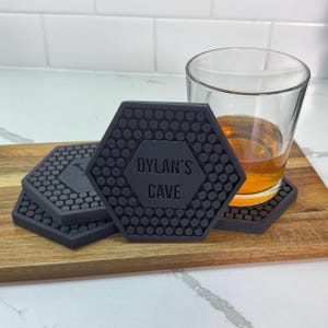 Può includere: Sottobicchieri esagonali neri con superficie strutturata e la scritta "DYLAN'S CAVE". Un bicchiere di liquido ambrato è appoggiato su un sottobicchiere. I sottobicchieri sono su una tavola di legno.
