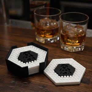 Puede incluir: Posavasos hexagonales en blanco y negro con un diseño central negro. Se ven dos vasos de líquido ámbar con cubitos de hielo. Los posavasos tienen una superficie texturizada y están apilados en un soporte.