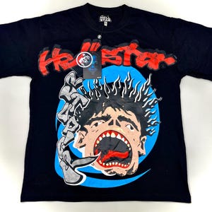 May include: Black t-shirt with a graphic design featuring the text "Hellstar" in red, a screaming face, and the word "STRAP" in gray. The design is set against a blue background. A tag is attached to the shirt.