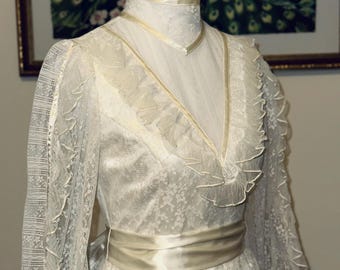 May include: A vintage ivory lace dress with long sleeves and a high neckline. The dress features a satin ribbon sash at the waist and delicate ruffle trim along the neckline and sleeves. The dress is displayed on a mannequin.