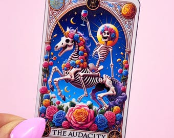 May include: A tarot card featuring a skeleton riding a unicorn, both adorned with flowers, against a blue background. The skeleton holds a wine glass. The card is framed with ornate details and the text "THE AUDACITY" at the bottom.