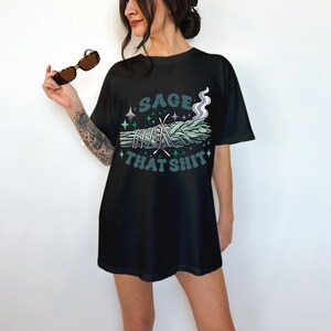 Puede incluir: Camiseta negra con un diseño gráfico que presenta un manojo de salvia con humo y el texto "SAGE THAT SHIT" en verde azulado y blanco. El diseño incluye estrellas y elementos en forma de cruz.