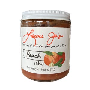 Puede incluir: Un tarro de salsa de melocotón con tapa blanca y etiqueta con texto "Lauri Jo's" y "Peach salsa". La etiqueta incluye ilustraciones de melocotones, tomates y un diente de ajo. Peso neto 227g.
