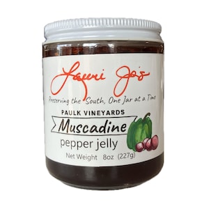 Puede incluir: Un tarro de gelatina de pimiento morrón Muscadine con tapa y etiqueta blancas. La etiqueta dice "Lauri Jo's" y "Preserving the South, One Jar at a Time". El tarro contiene gelatina oscura y está etiquetado "Peso neto 227g (8oz)."