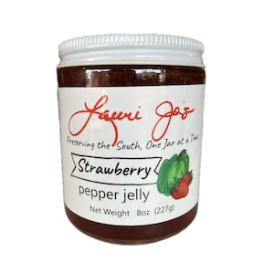 Puede incluir: Un tarro de gelatina de fresa y pimiento con tapa blanca y etiqueta con texto "Laurie Jo's" y "Strawberry pepper jelly". La etiqueta incluye un gráfico de pimientos y fresas. Peso neto 227g.