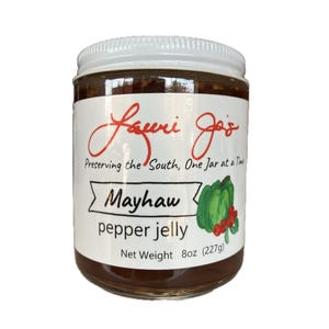 Puede incluir: Un tarro de cristal de gelatina de pimiento Mayhaw con tapa blanca. La etiqueta dice "Lauri Jo's" y "Preserving the South, One Jar at a Time". La etiqueta también presenta un pimiento verde y bayas rojas. Peso neto 227g.