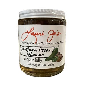 Puede incluir: Un tarro de cristal de gelatina de pimiento jalapeño y nuez pecana sureña con tapa blanca. La etiqueta dice "Laurie Jo's" y "Preservando el Sur, un tarro a la vez". El tarro contiene 227g de gelatina.