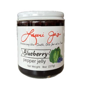 Puede incluir: Un tarro de mermelada de arándanos y pimientos con tapa blanca. La etiqueta dice "Lauri Jo's" y "Blueberry pepper jelly". El tarro contiene una gelatina de color púrpura oscuro. El peso neto es de 227g.