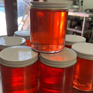 Puede incluir: Una colección de frascos de vidrio transparente llenos de un líquido rojo translúcido vibrante. Cada frasco está coronado con una tapa plateada. Los frascos están apilados, creando una disposición visualmente atractiva.