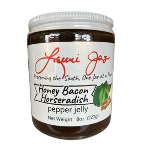 Puede incluir: Un tarro de gelatina de pimienta, miel, tocino y rábano picante con tapa y etiqueta blancas. La etiqueta dice "Lauri Jo's" y "Preserving the South, One Jar at a Time." El peso neto es de 227g.