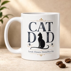 Puede incluir: Una taza de cerámica blanca con el texto CAT DAD en letras grandes de color azul oscuro, con una silueta de gato negro que reemplaza la letra A. Debajo del gato, el texto dice Loved. Chosen. Owned by a Cat. Adornos dorados enmarcan el diseño.
