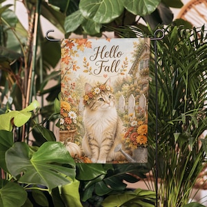 Puede incluir: Una bandera de jardín rectangular con un gato atigrado marrón que lleva una corona de flores, sentado en un jardín con calabazas, flores y una regadera. El texto Hello Fall está impreso en cursiva marrón en la parte superior.