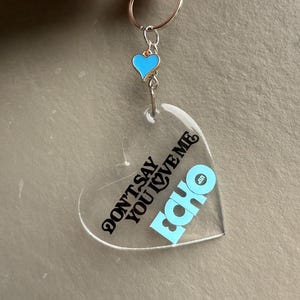 May include: Clear heart-shaped keychain with the text "DON'T SAY YOU LOVE ME" and the word "ECHO" in light blue. A small blue heart charm with a gold border hangs above the heart. The keychain has a silver ring.