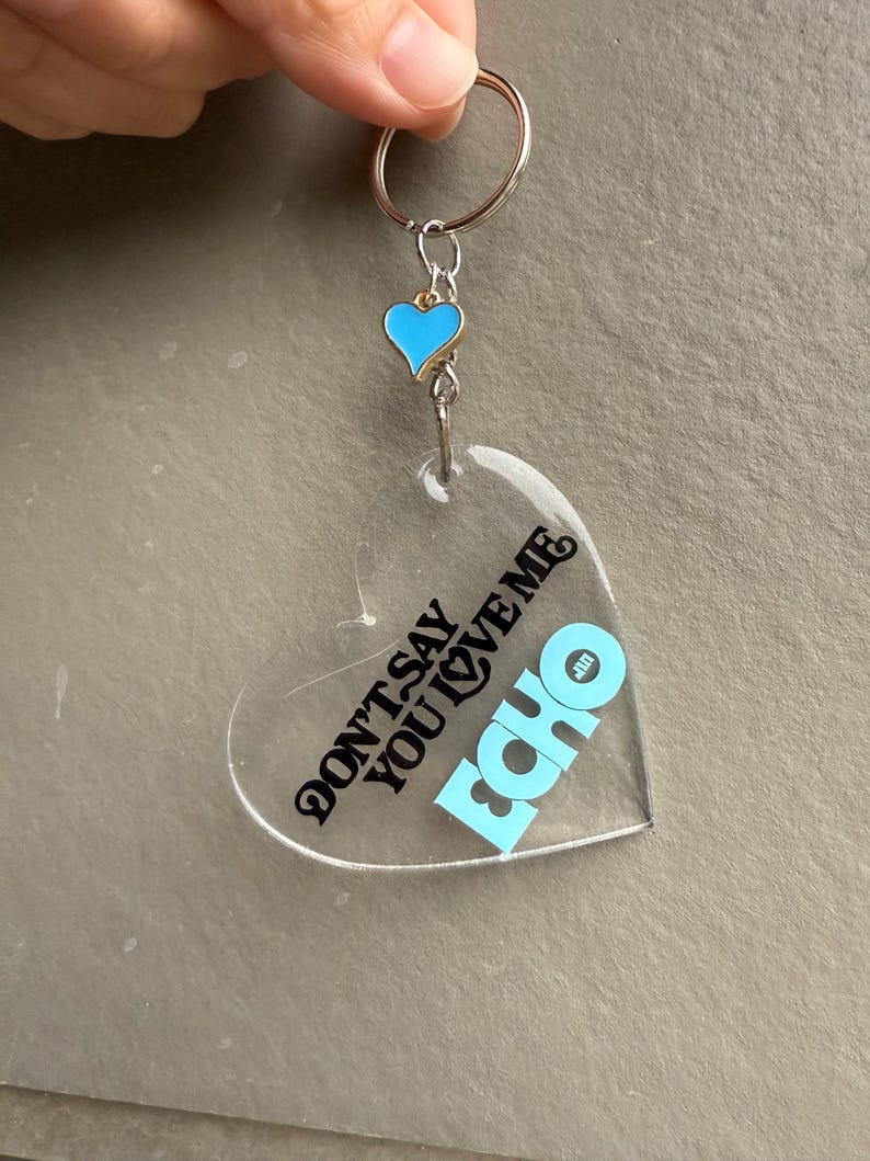 May include: Clear heart-shaped keychain with the text "DON'T SAY YOU LOVE ME" and the word "ECHO" in light blue. A small blue heart charm with a gold border hangs above the heart. The keychain has a silver ring.