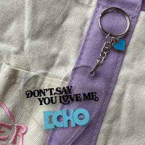 May include: A clear heart-shaped keychain with the text "DON'T SAY YOU LOVE ME ECHO" in black and blue. The keychain is attached to a silver keyring and a blue heart charm. The background includes a canvas tote bag with purple accents.
