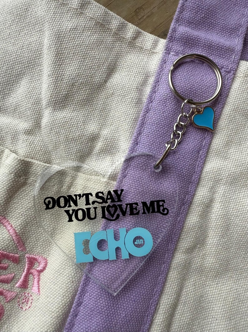 May include: A clear heart-shaped keychain with the text "DON'T SAY YOU LOVE ME ECHO" in black and blue. The keychain is attached to a silver keyring and a blue heart charm. The background includes a canvas tote bag with purple accents.