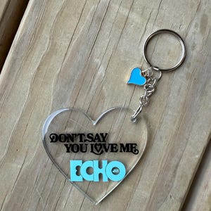 Peut inclure: Porte-clés transparent en forme de cœur avec le texte "DON'T SAY YOU LOVE ME ECHO" en noir et bleu clair. Un petit cœur bleu clair et un porte-clés argenté complètent le design. Le porte-clés est sur une surface en bois.