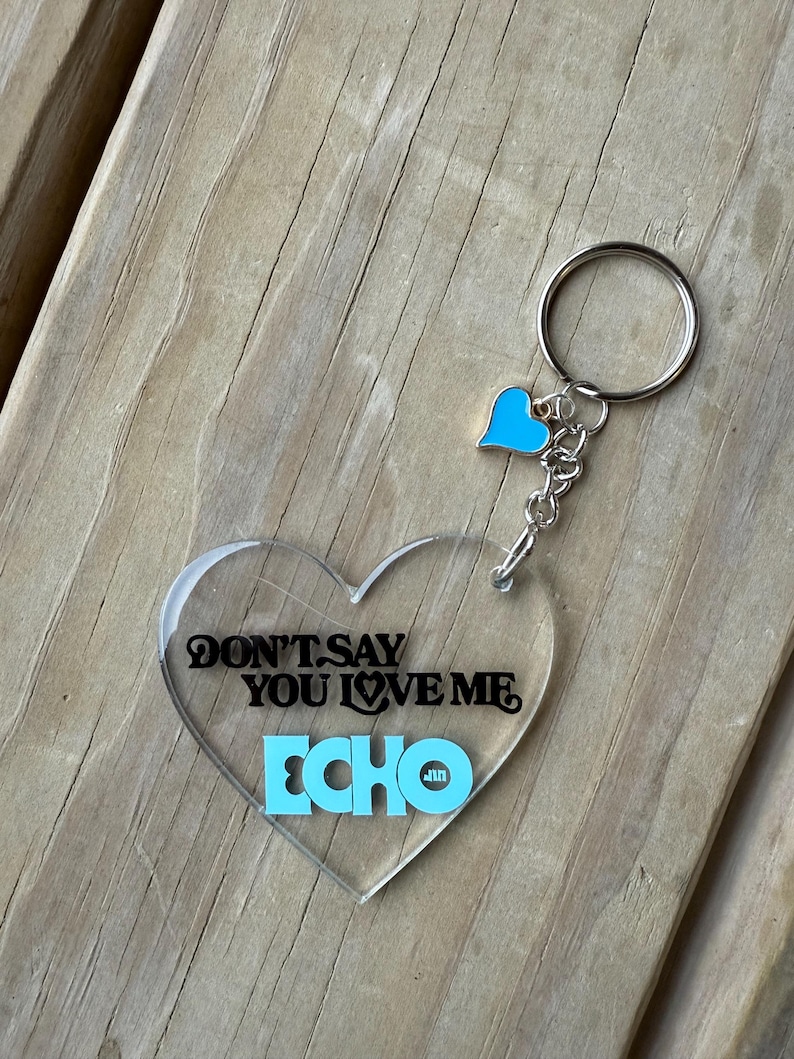 May include: Clear heart-shaped keychain with the text "DON'T SAY YOU LOVE ME ECHO" in black and light blue. A small light blue heart charm and a silver keyring complete the design. The keychain is on a wooden surface.