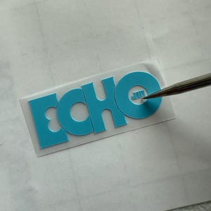 May include: A teal-colored heat transfer vinyl with the word "ECHO" cut out. The letters are bold and sans-serif. A tool is positioned near the letter "O". The vinyl is on a white surface.