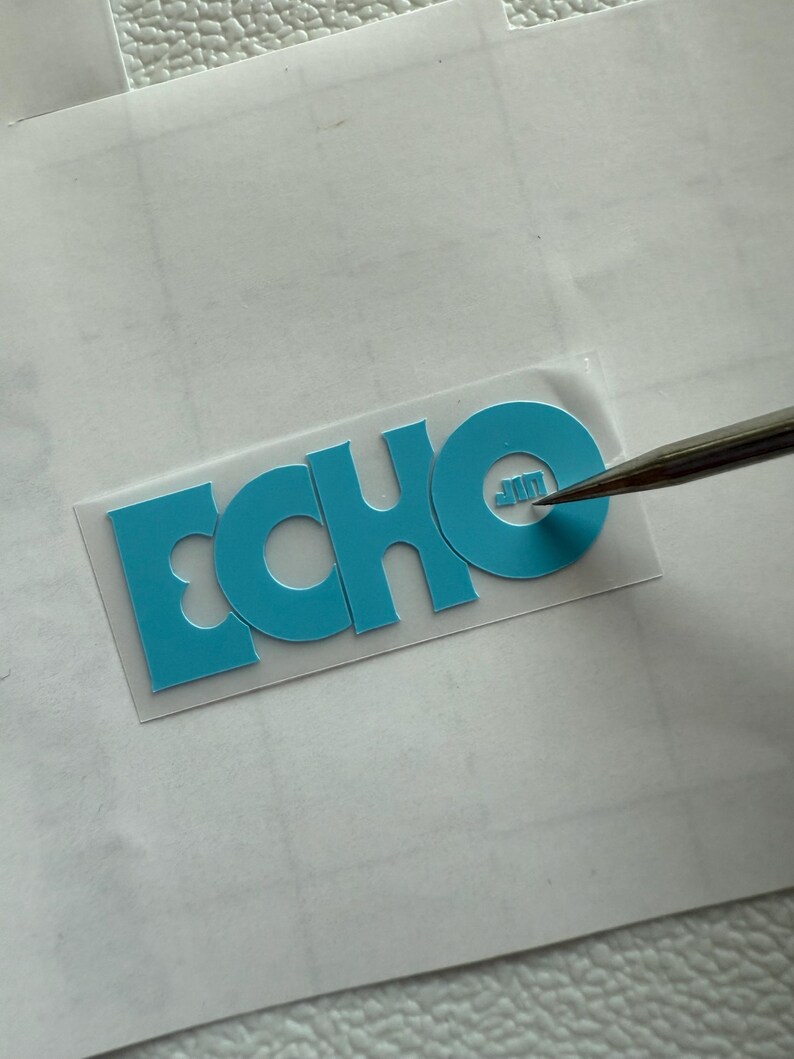 May include: A teal-colored heat transfer vinyl with the word "ECHO" cut out. The letters are bold and sans-serif. A tool is positioned near the letter "O". The vinyl is on a white surface.