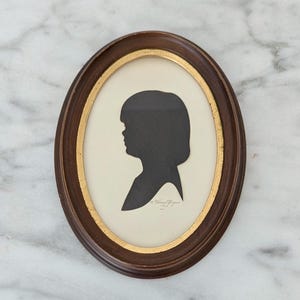 May include: Oval framed silhouette portrait of a person in profile, with a dark brown wooden frame and a gold inner border. The silhouette is black against a cream-colored background. The frame is resting on a white marble surface.