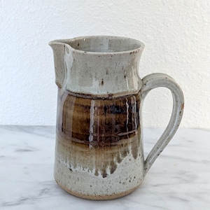 May include: A handmade ceramic pitcher with a handle and spout. The pitcher features a two-tone design, with a light gray top and a brown base. The pitcher is approximately 8 inches tall and is suitable for serving beverages.