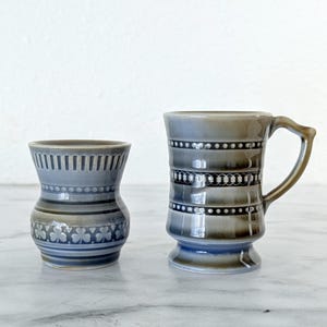 May include: Two ceramic pieces: a small vase and a mug. The vase is blue-grey with vertical lines and clover patterns. The mug is blue-grey with a handle and horizontal bands of dots and lines. Both have a vintage aesthetic.
