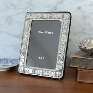 May include: Silver-plated photo frame with a decorative floral border. The frame is rectangular and holds a 5x7 inch photo. The frame is displayed on a wooden surface, with a marble background. The text "Silver Plated" and "5x7" are visible on the frame.