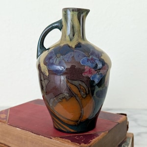 May include: A small, decorative ceramic pitcher with a handle, featuring a floral design in shades of blue, brown, and orange. The pitcher is set atop two antique books with red and tan covers. The background is a plain white.