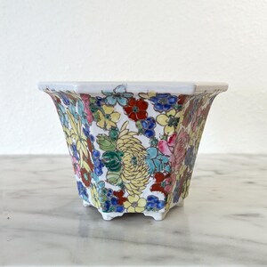 May include: A square, white ceramic planter with a vibrant floral pattern. The design features a variety of colorful flowers, including red, yellow, blue, and pink blooms, set against a white background. The planter has a slightly flared shape and small feet.