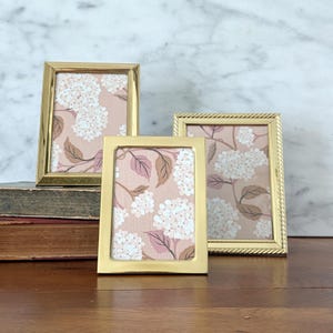 May include: Three gold-colored picture frames of varying sizes. Each frame holds a floral print with white hydrangea blooms, pink accents, and brown leaves against a soft pink background. The frames are displayed on a wooden surface.