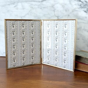 May include: A gold-toned, double-hinged picture frame. The frame holds two rectangular panels with a floral and striped pattern in shades of gray and cream. The frame is open, revealing the patterned interior.