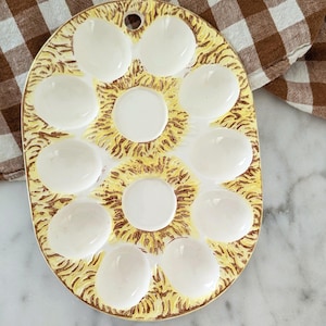 May include: Oval ceramic egg serving dish with 12 white egg-shaped indentations. The dish has a yellow and brown floral design and a gold rim. A small hole is at the top for hanging. The dish is on a marble surface.