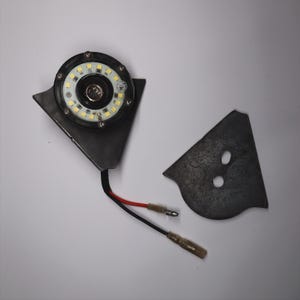 May include: A circular LED light with a black metal housing and a black wire with red and black wires attached. The light is mounted on a dark gray metal bracket, with a separate matching bracket piece.