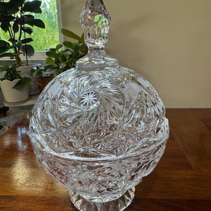 May include: A clear, spherical crystal candy dish with a matching lid featuring a pointed finial. The glass surface is intricately cut with a repeating starburst and cross-hatch pattern, resting on a circular, faceted pedestal base.