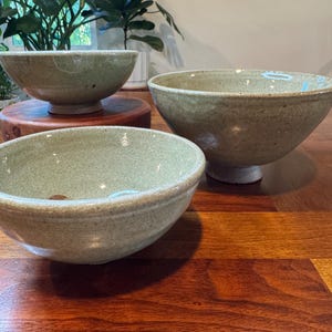 May include: Three ceramic bowls with a speckled light green glaze. Two bowls feature a raised pedestal base, while the third bowl has a rounded bottom. The surfaces have a subtle texture with visible variations in the glaze color and finish.