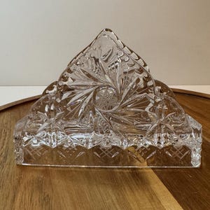 May include: A clear, cut-glass napkin holder with a triangular top and a rectangular base. The glass is intricately patterned with starburst and diamond designs. The holder is designed to hold napkins upright.