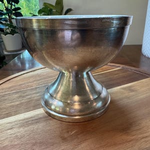 May include: A silver-toned metal pedestal bowl with a smooth, rounded basin and a flared base. The bowl features a polished, reflective surface with subtle variations in tone, showcasing a wide, open top and a sturdy, elevated stem design.