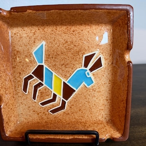 May include: A square ceramic ashtray with a speckled tan and brown glaze. The center features a stylized geometric dog graphic composed of blue, yellow, and brown segments outlined in white, set against the textured, mottled tan background.